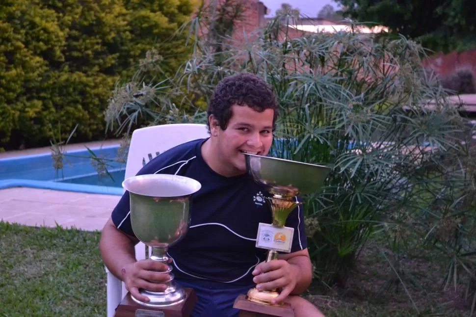 CAMPEÓN EN TODOS LADOS. Santiago Iglesias Valdez posa en el fondo de su casa a lo  Rafa Nadal, mordiendo el trofeo obtenido el domingo en Río Cuarto. Del otro lado, la copa del Argentino 2011.  