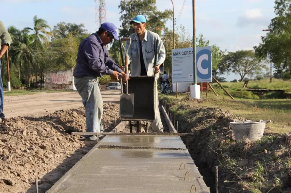 EN EJECUCIÓN. Obreros colocan mezcla en una vereda de Río Colorado; detrás se lee el cartel “Más Cerca”. COMUNICACÍON PÚBLICA (archivo)