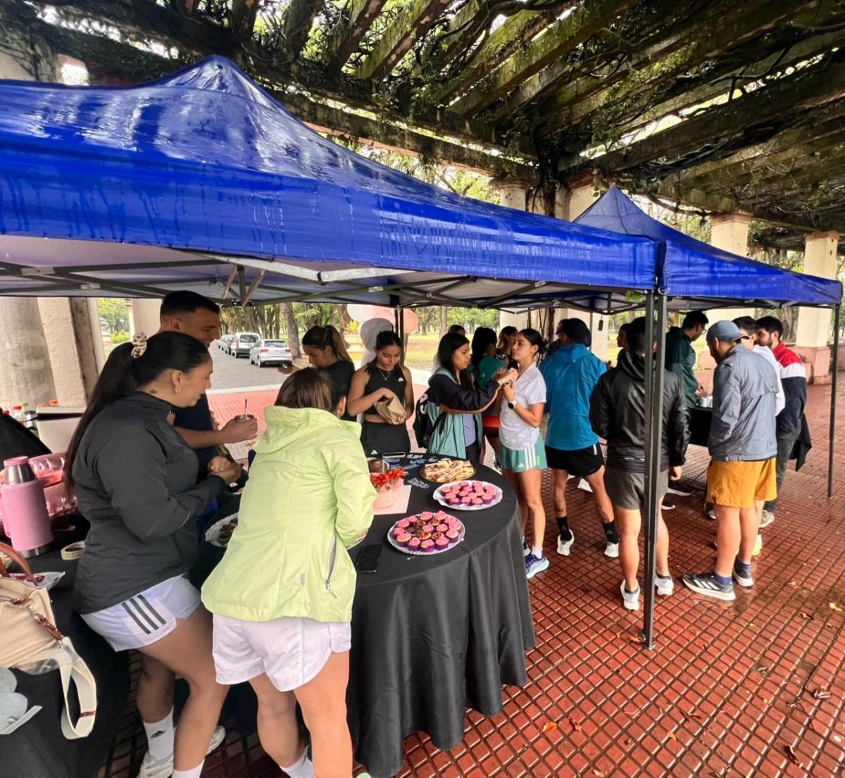 Bajo el gazebo, la lluvia se volvió apenas un murmullo. Sobre los tablones improvisados como mesas, se desplegaban tortas caseras, alfajores de maicena, frutas cortadas y termos con mate y café.
