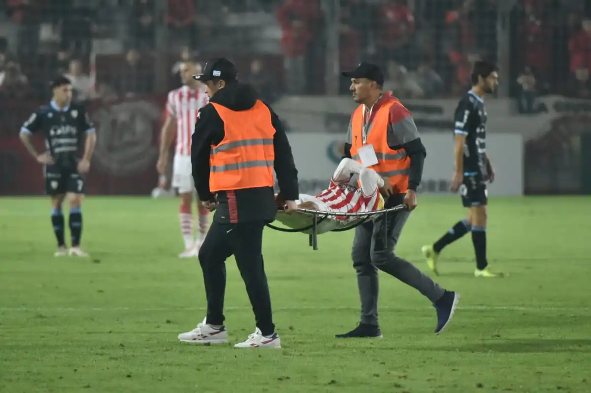 ALERTA. Dos asistentes trasladan en camilla a Kevin López, con la pierna inmovilizada y el gesto serio, mientras el resto de los jugadores observa en silencio y el partido queda en segundo plano.